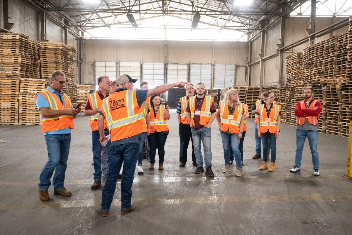 Group of 48forty employees wearing safety vests on pallet facility plant floor. Stacks of recycled white wood pallets are in the background.